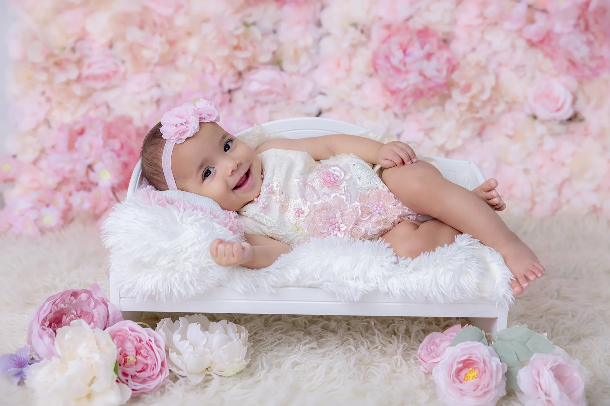 A smiling baby girl lying on a white fluffy blanket on a small white bed, surrounded by pink and white flowers, with a pink flower headband and floral dress, set against a background of pink flower wall.
