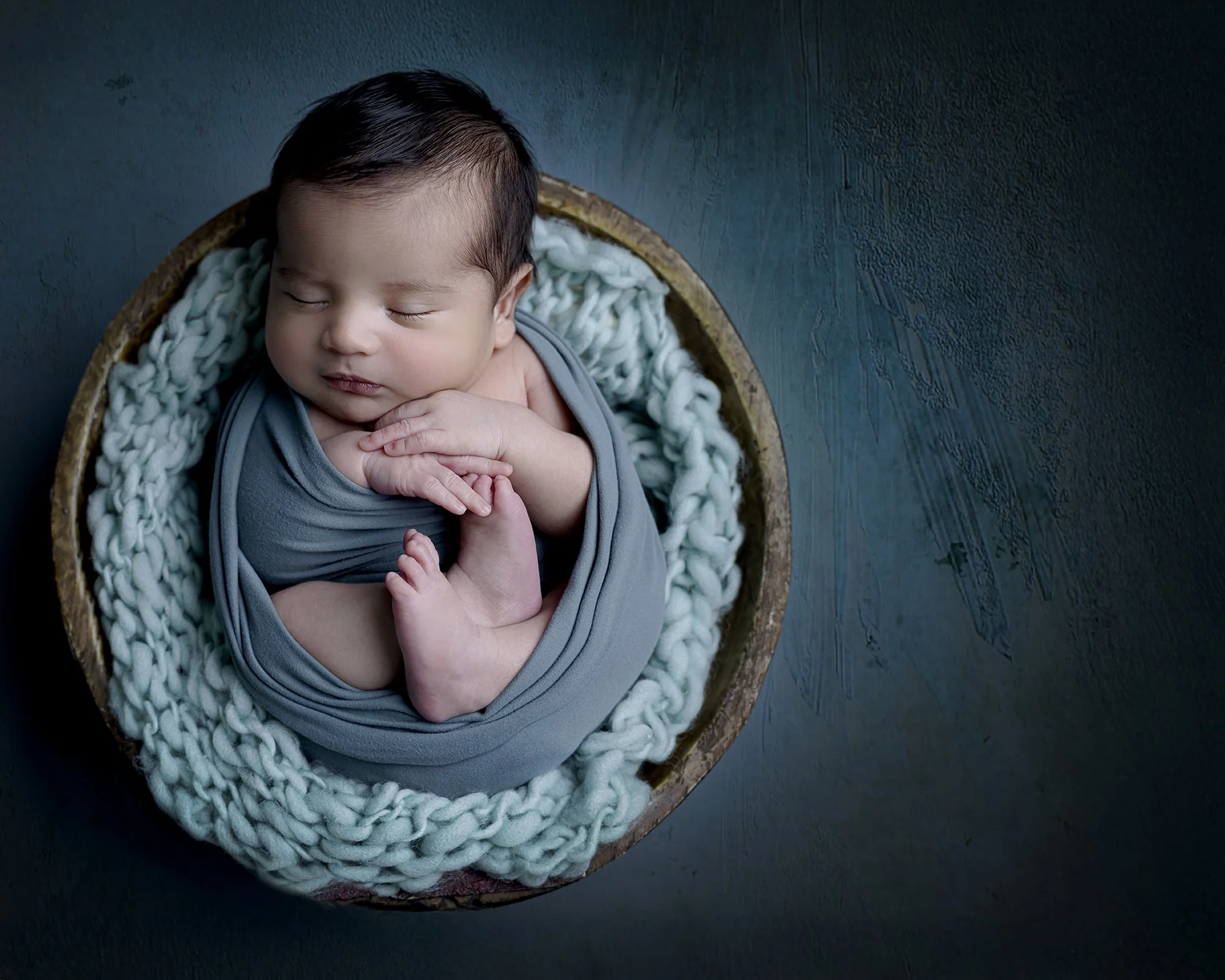 A sleeping newborn baby wrapped in a gray blanket, resting in a cozy round basket lined with chunky knit fabric, on a dark surface.