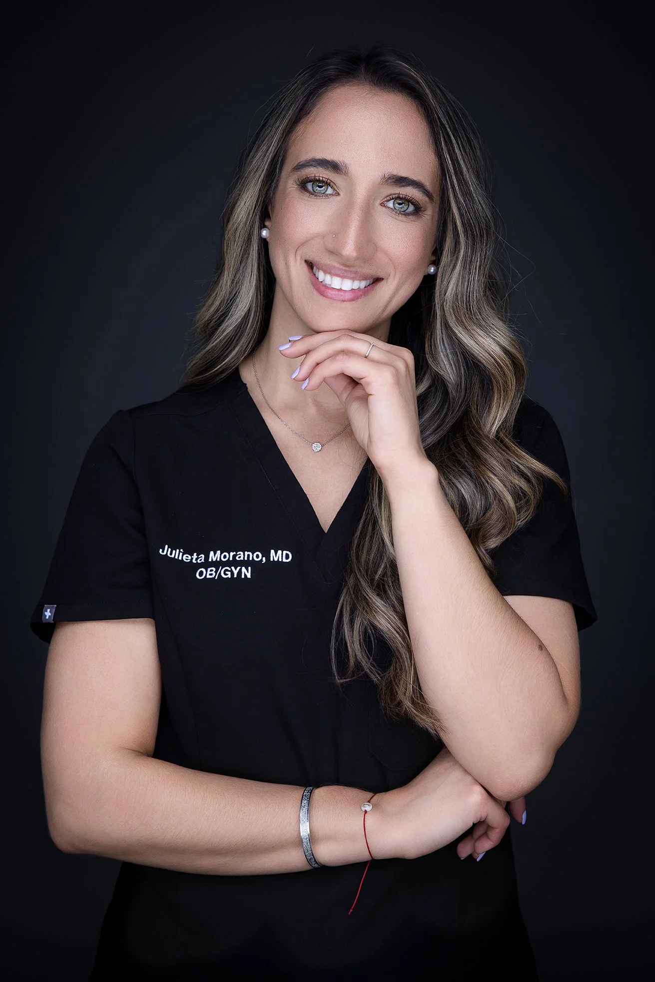 A smiling woman with long wavy hair, wearing a black medical uniform with 'Julieta Morano, MD OB/GYN' embroidered on it, posing against a dark background.