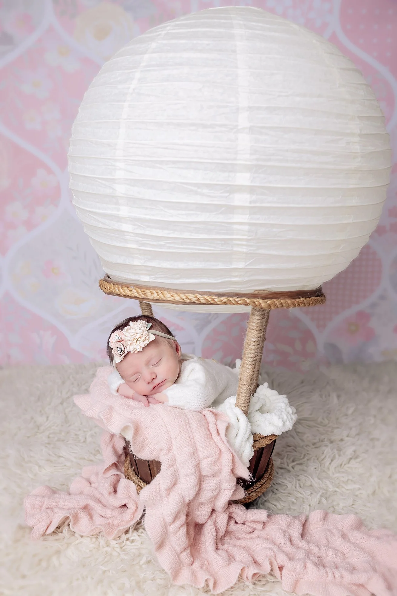 A sleeping baby girl with a floral headband, resting on a pink and white blanket inside a miniature hot air balloon basket with a white paper lantern as the balloon, on a soft cream-colored surface.