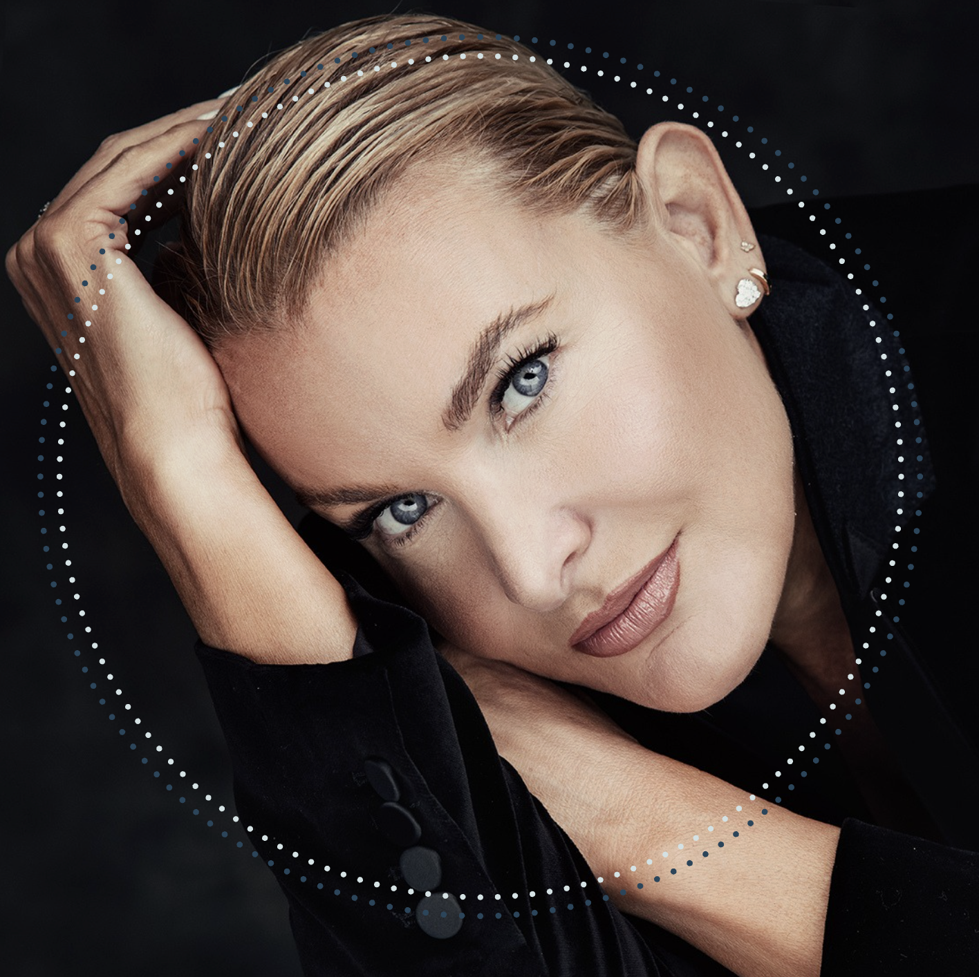 Close-up portrait of a woman with blonde hair, blue eyes, and fair skin, resting her head on her hand against a black background, wearing a black top and jewelry.