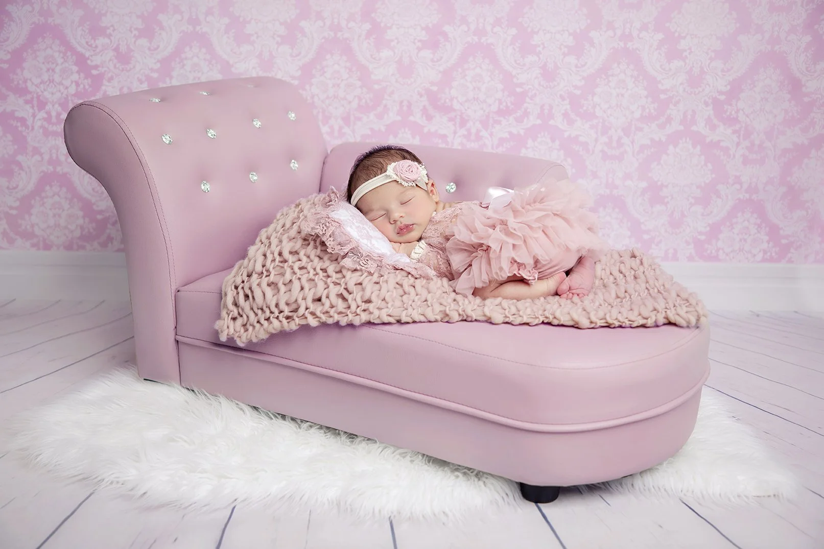 A sleeping baby girl on a pink sofa with ruffled pink dress and headband, surrounded by pink and white decor.