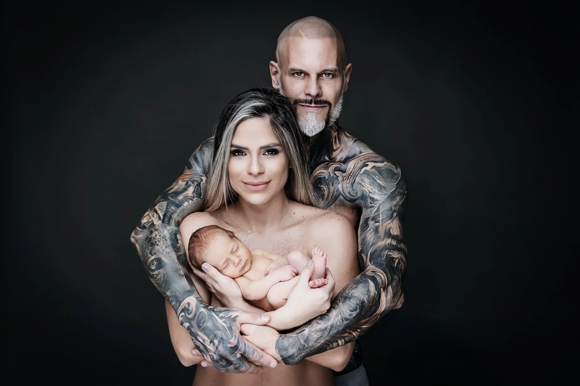 A tattooed man and woman holding a sleeping newborn baby against a dark background.