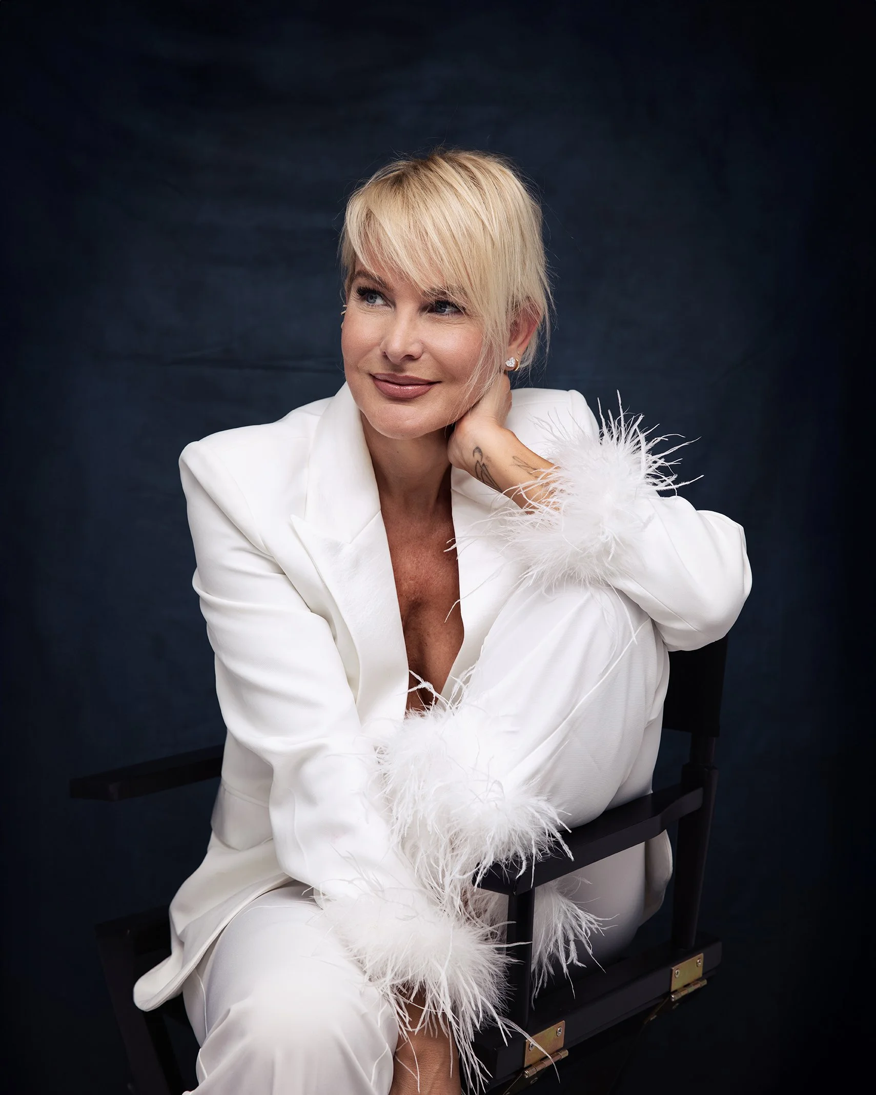 A blonde woman in a white suit with feathered cuffs sitting on a black chair against a dark background.