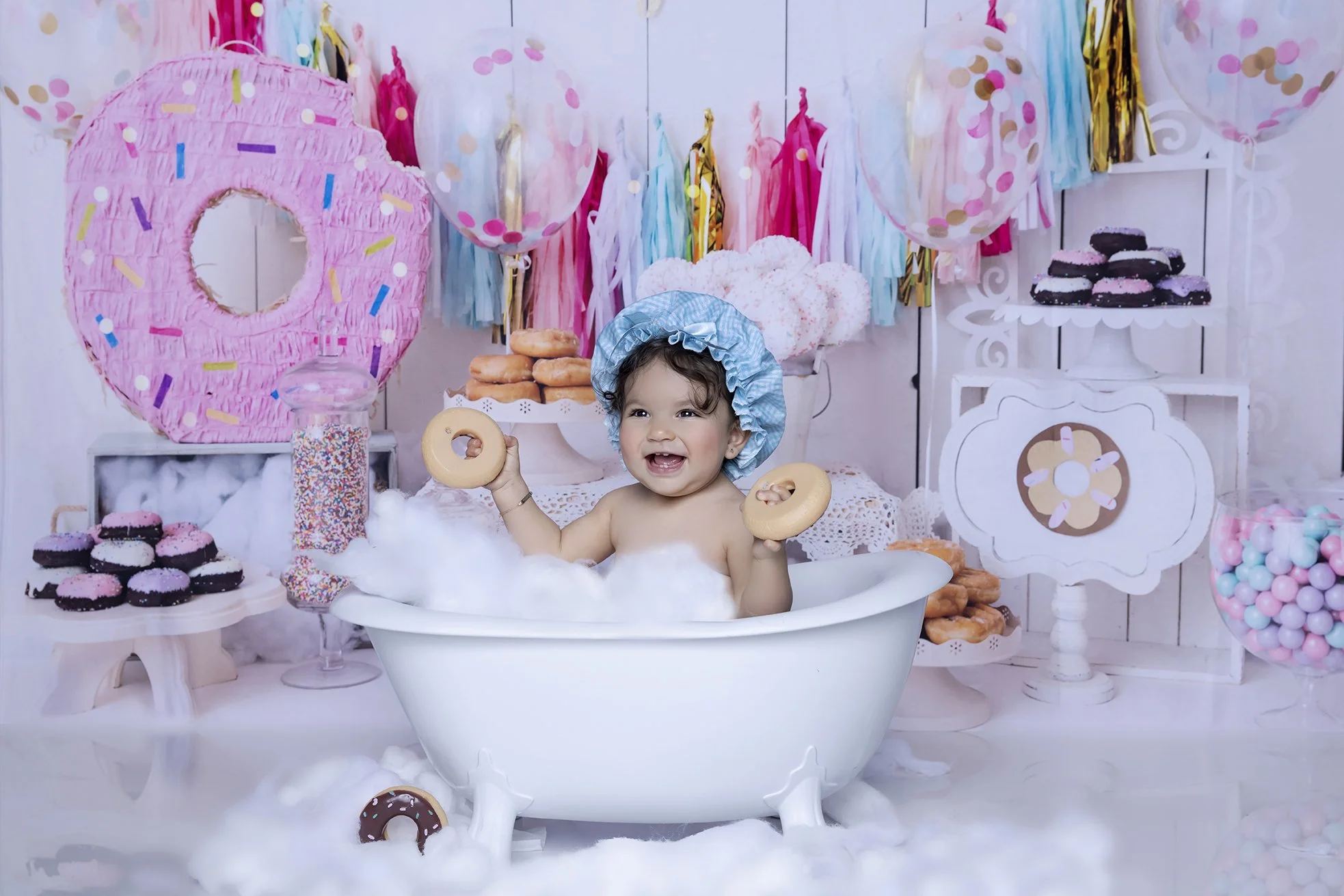 A young child smiling in a bathtub filled with bubbles, holding two donuts, wearing a blue shower cap, with a colorful birthday party setup in the background, including balloons, donuts, and decorations.