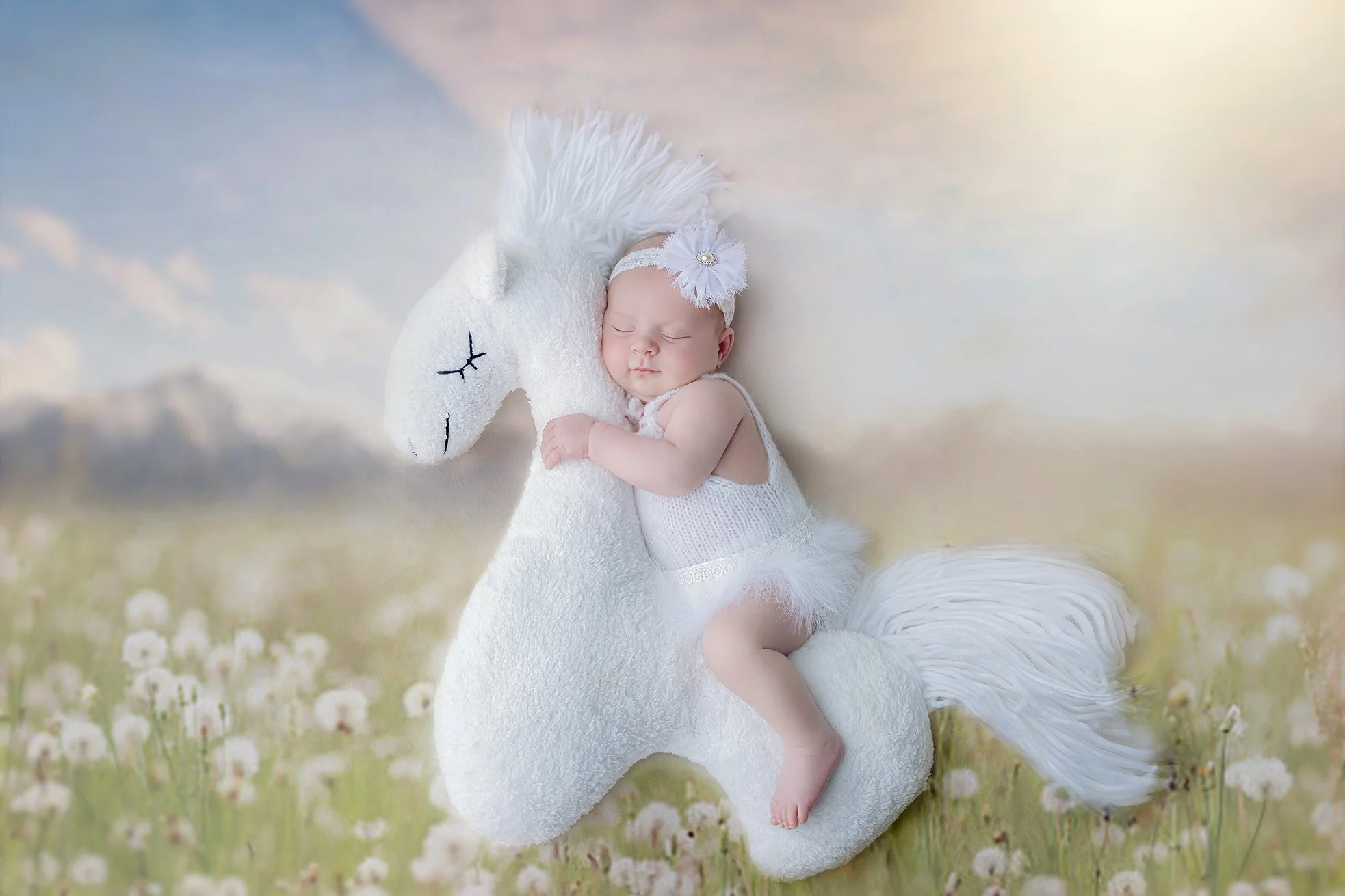 A sleeping baby girl in a white dress and headband with a flower, cuddling a plush white horse stuffed animal in a dreamy outdoor field with soft sunlight.