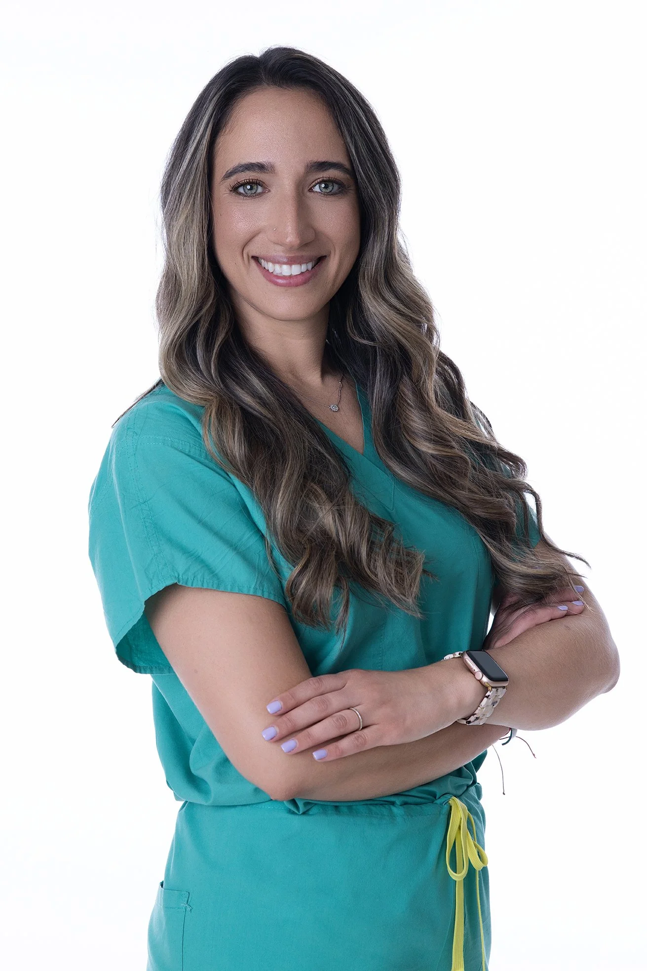 A smiling woman with long wavy brown hair, wearing teal medical scrubs and a smartwatch, standing with arms crossed against a white background.