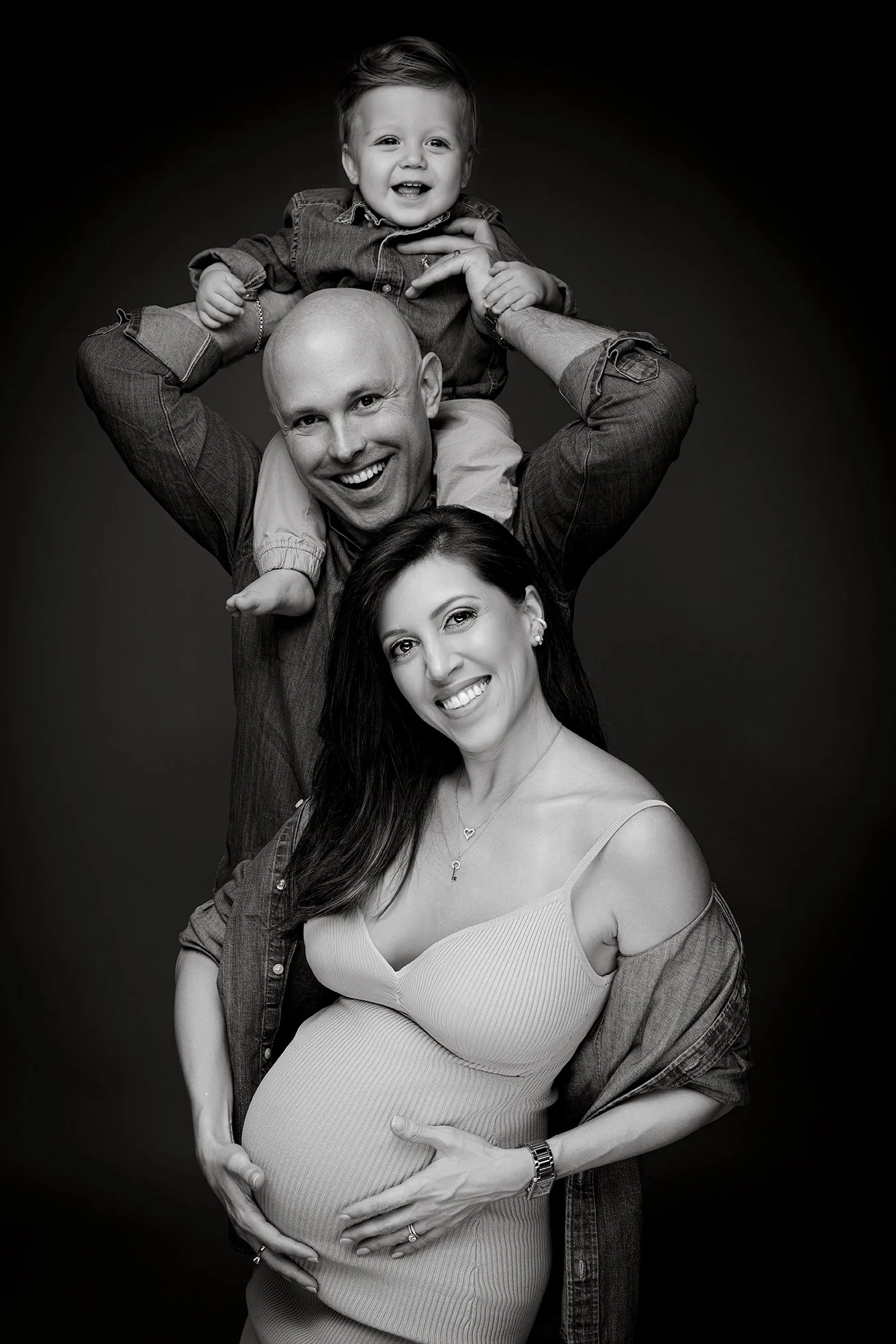 Black and white photo of a happy family of four, with the pregnant mother in the front, the father behind her, and their young child sitting on the father's shoulders, all smiling at the camera.