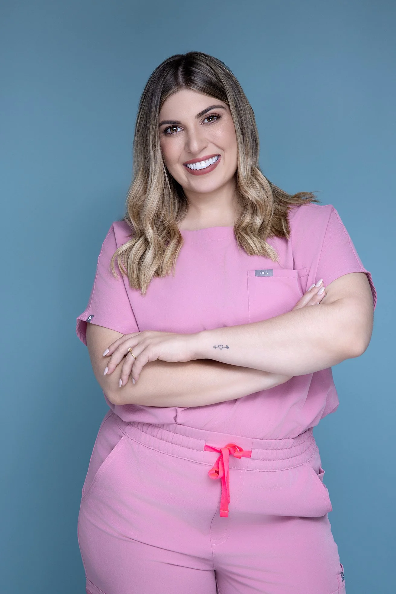 A young woman with shoulder-length wavy hair, wearing a pink medical scrub top and matching scrubs pants, smiling with arms crossed against a blue background.