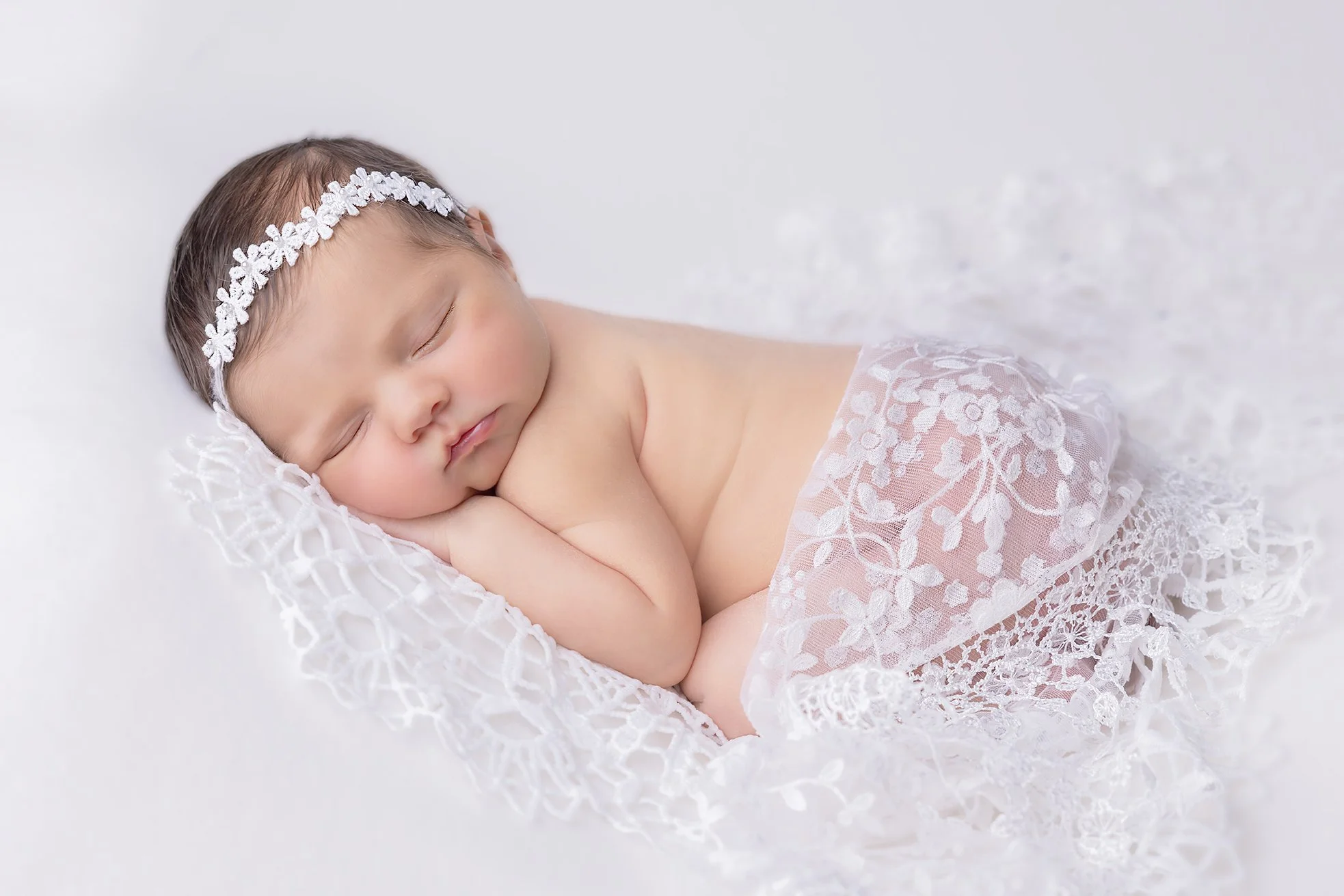A sleeping baby girl with a white floral headband, wrapped in a sheer lace fabric, resting on a soft white surface.