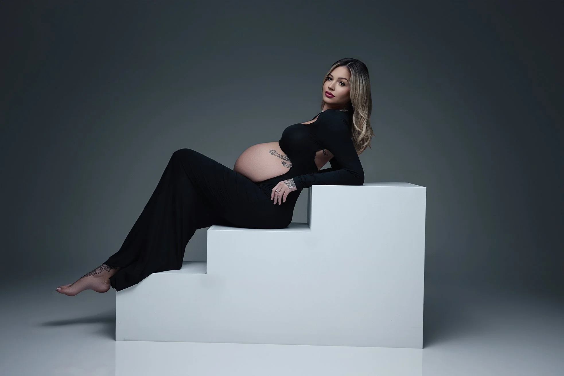 Pregnant woman in black dress lying on white stepped platform against gray background.