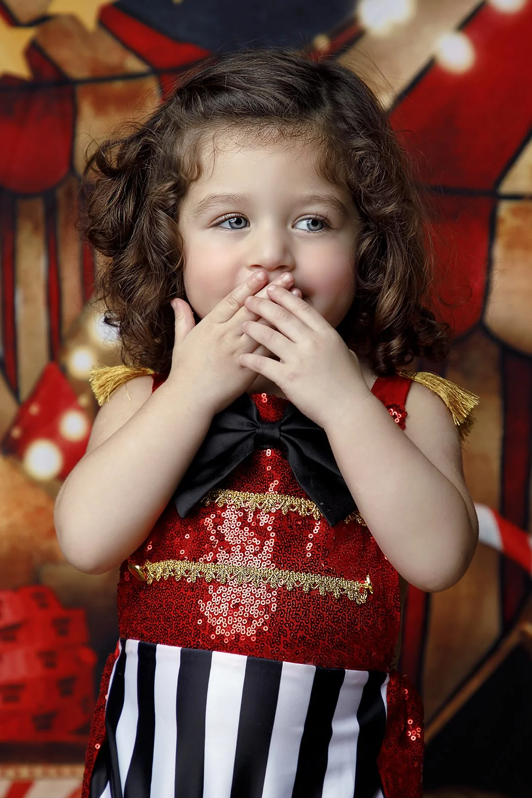 Young girl with curly hair covering her mouth with her hands, dressed in a red sequined dress with gold trim and black bow, standing against a festive background with Christmas decorations.