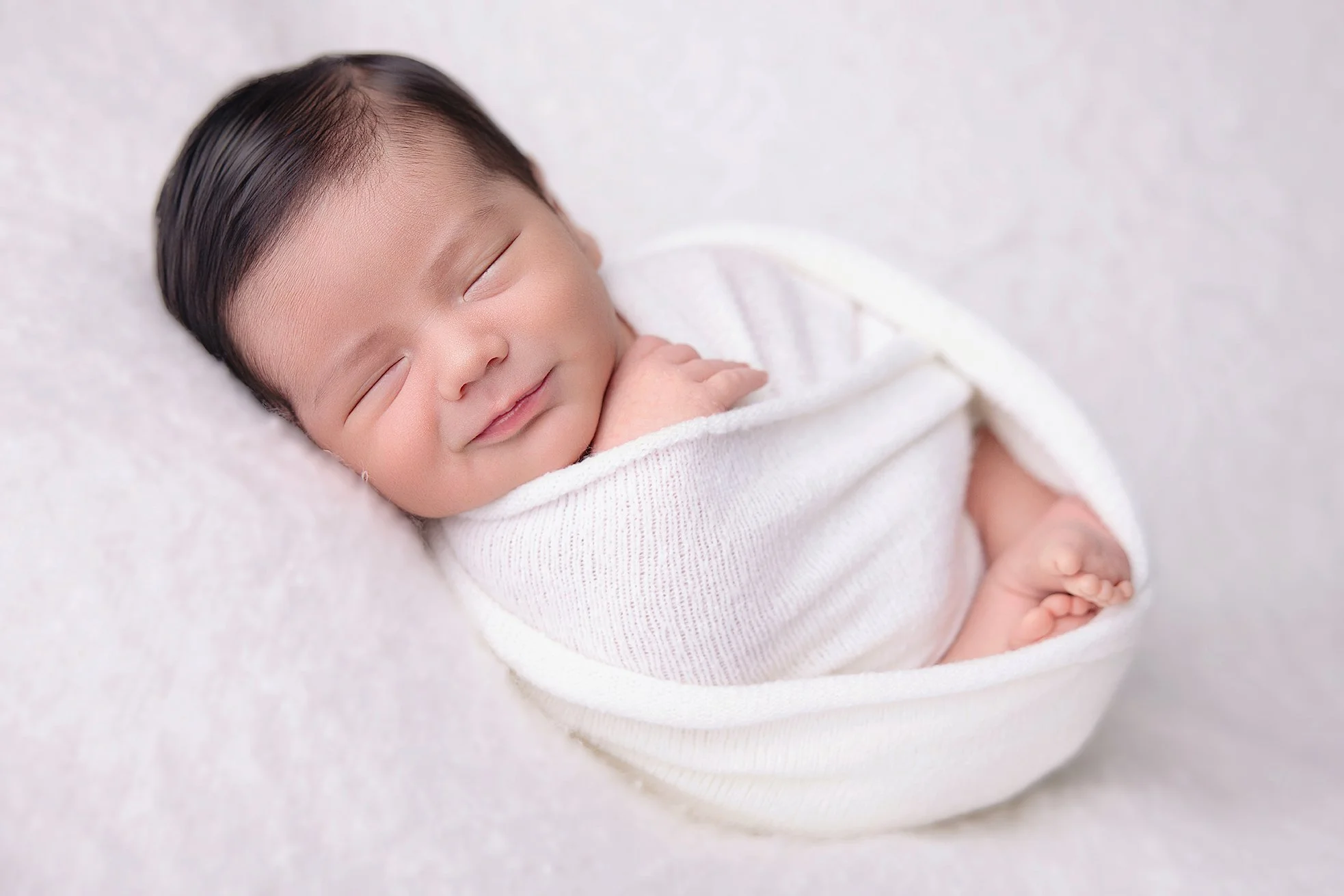 Close-up of a smiling newborn baby with dark hair, wrapped in a white blanket, lying on a soft light-colored surface.