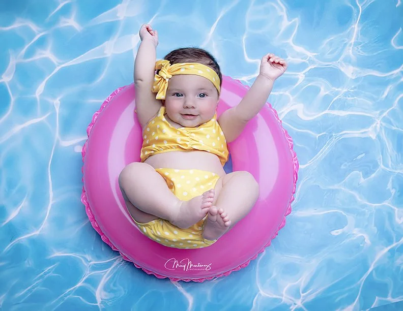 A smiling baby girl floating on a pink inner tube in a swimming pool, wearing a yellow polka dot swimsuit and matching headband.