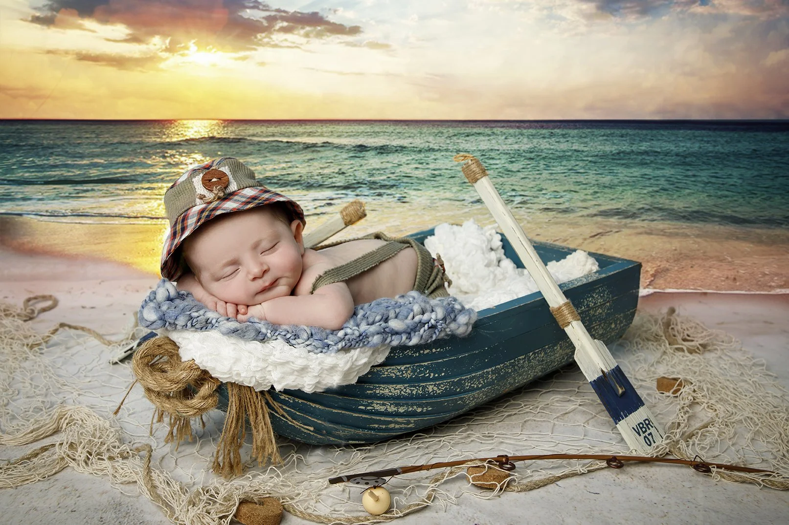 A sleeping baby lies in a small boat filled with white fluffy material, on a beach with a sunset over the ocean in the background. The baby wears a fisherman hat and a suspenders shirt, and is resting on a blanket. A small oar with the markings 'VBR 