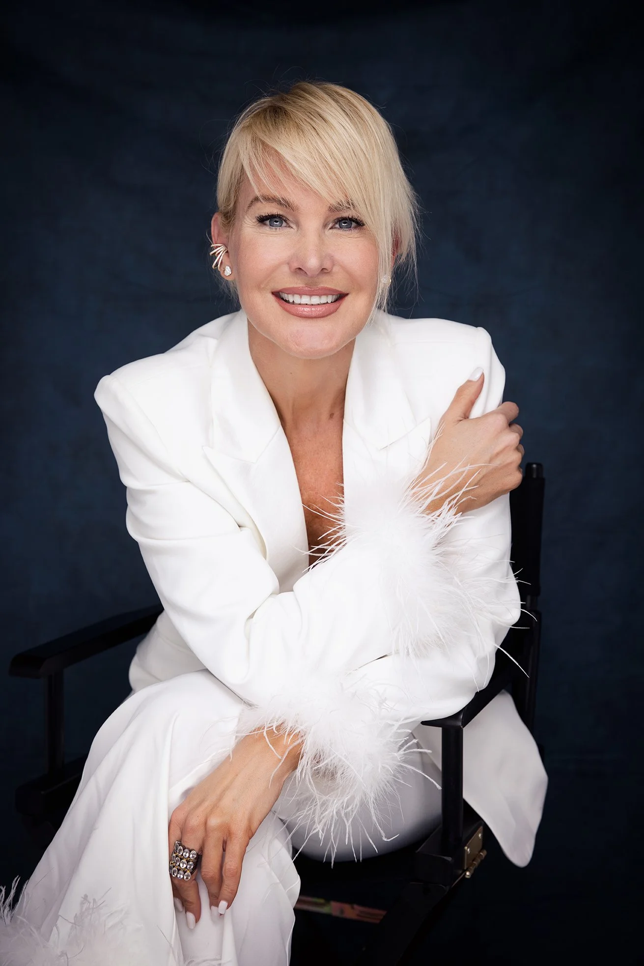 A smiling woman with short blonde hair, dressed in a white suit with feathered cuffs, sitting on a black director's chair against a dark background.