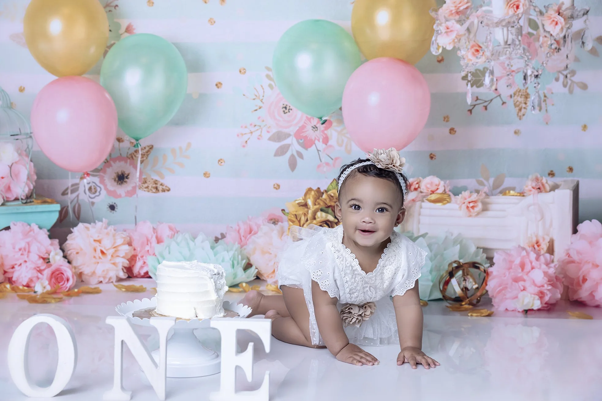 A baby girl in a white dress with lace details and a headband with a flower, on a decorated birthday backdrop. There are pink, green, and yellow balloons, pink and peach flowers, a small cake, and large white letters spelling 'ONE' in front.