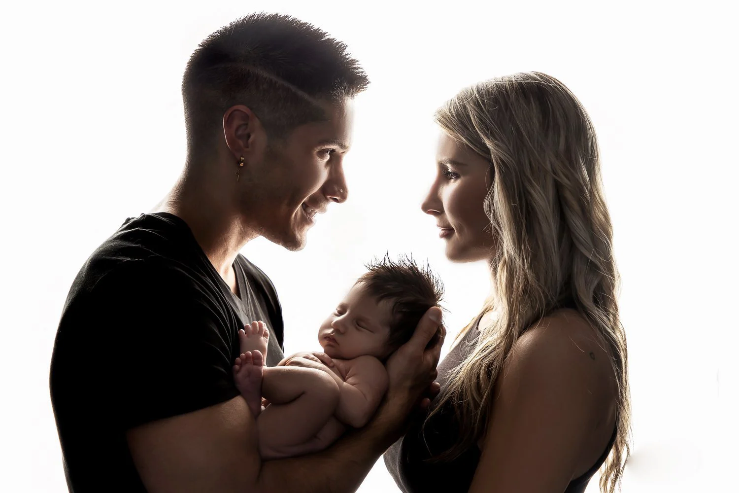 A happy couple holding their newborn baby close to each other, smiling and gazing into each other's eyes, with a backlit white background.