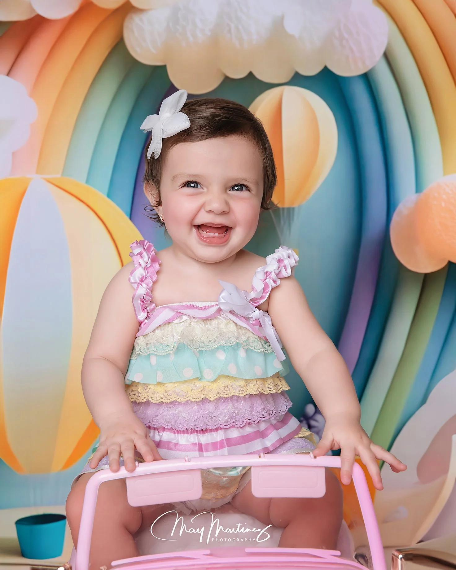 A smiling young girl with short brown hair, a white bow, wearing a colorful ruffled dress, seated in front of a rainbow and cloud backdrop.
