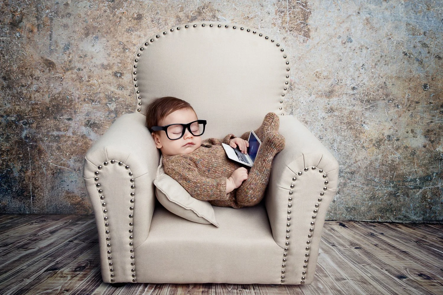A young child with glasses sleeping on an armchair while holding a small laptop, with a pillow under their head, in a room with a textured wall and wooden floor.
