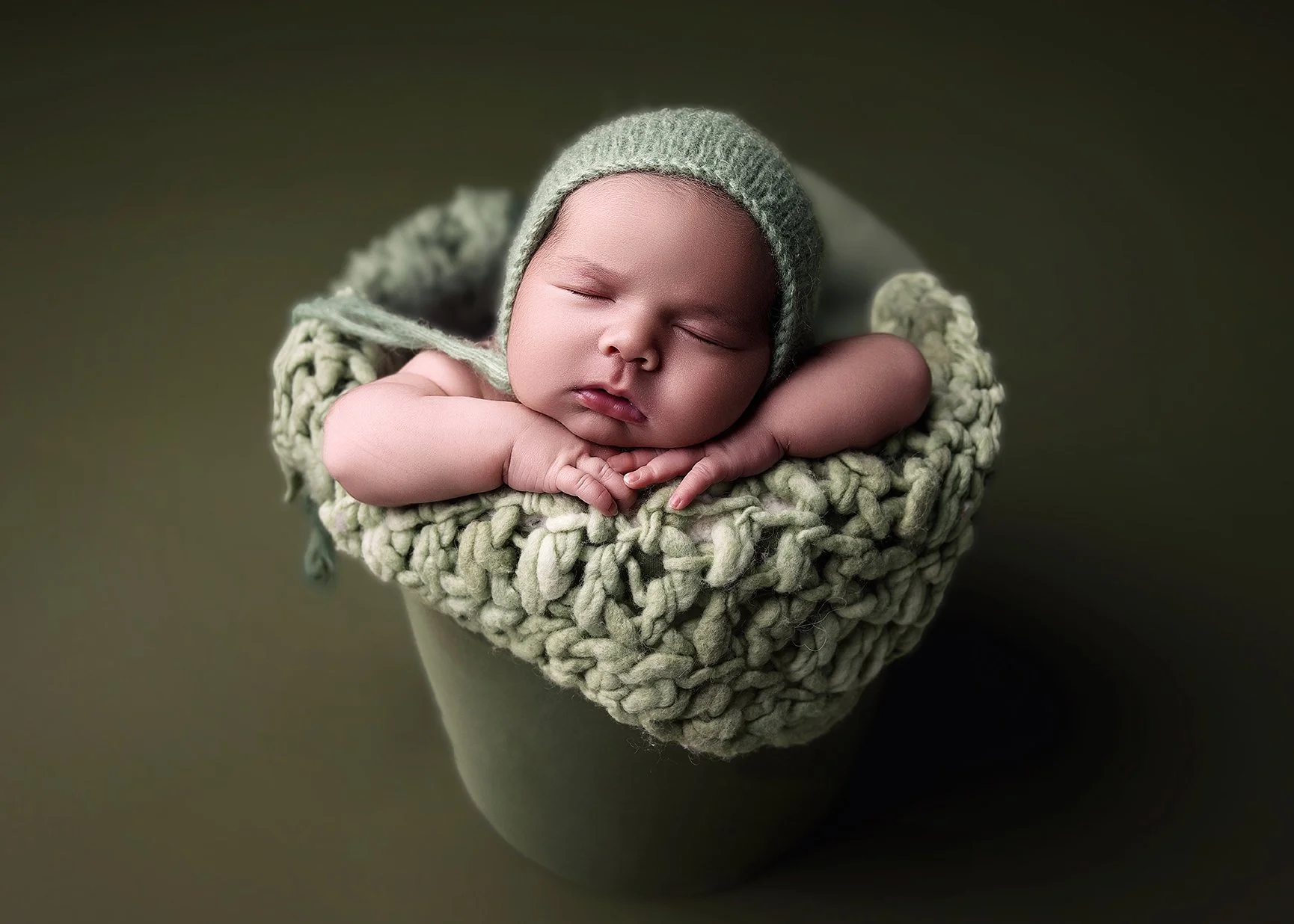 A sleeping baby inside a knitted green hat, resting on a fluffy green blanket in a cup.