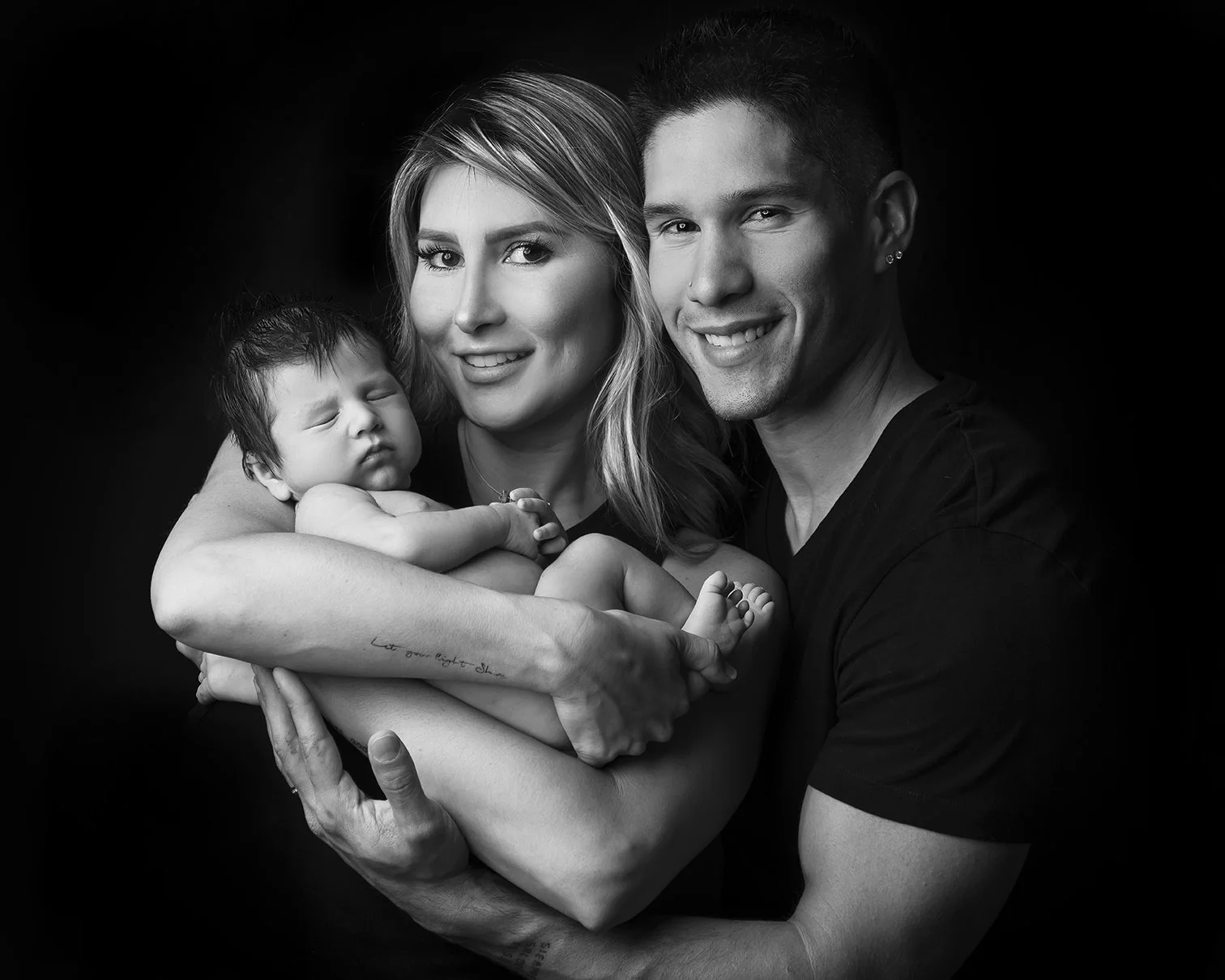 A family black and white portrait featuring two adults and a newborn baby. The woman is holding the baby close, and the man is standing beside her, all smiling gently.