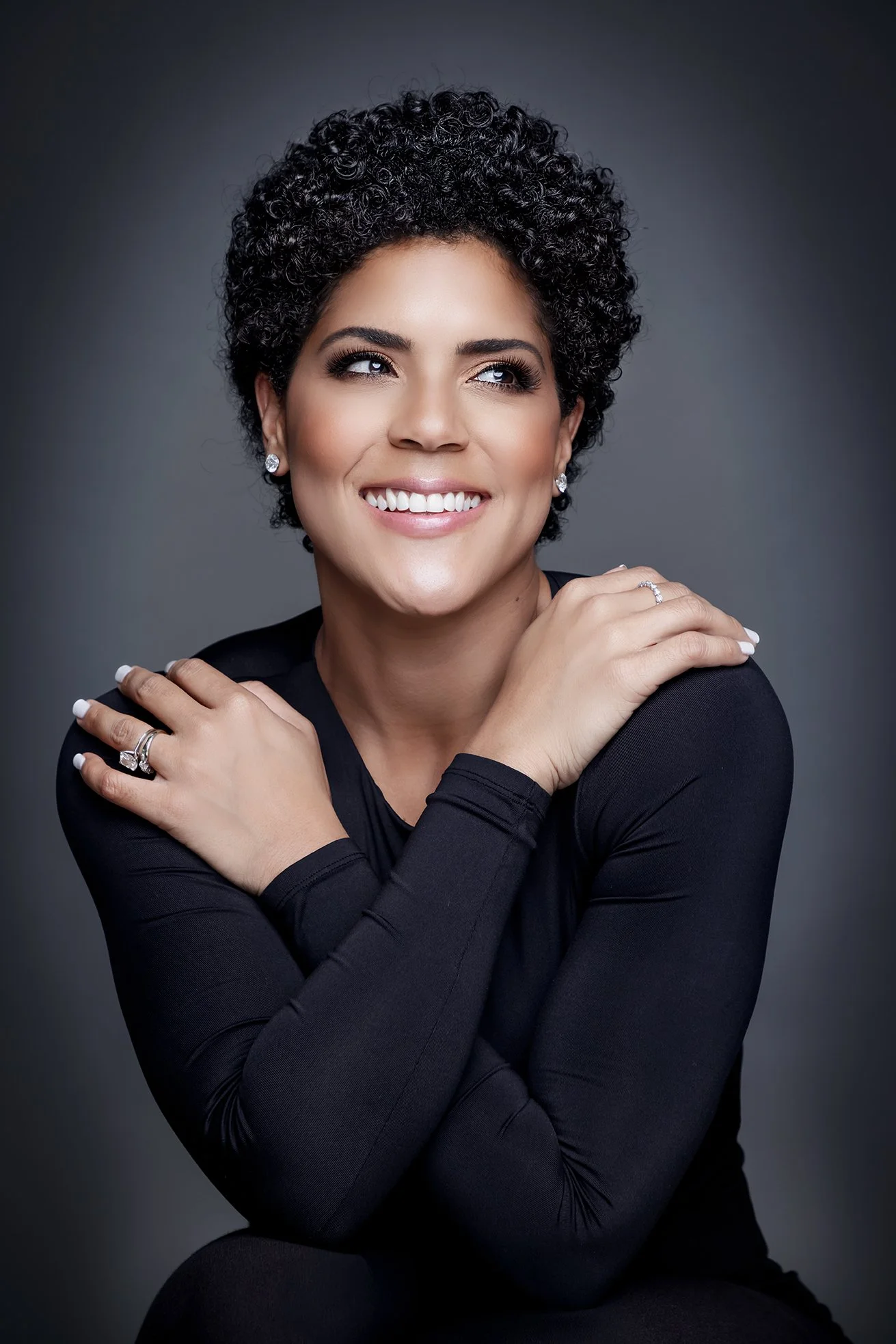 Portrait of a woman with curly hair, smiling, wearing a black top, and showing jewelry, against a gray background.