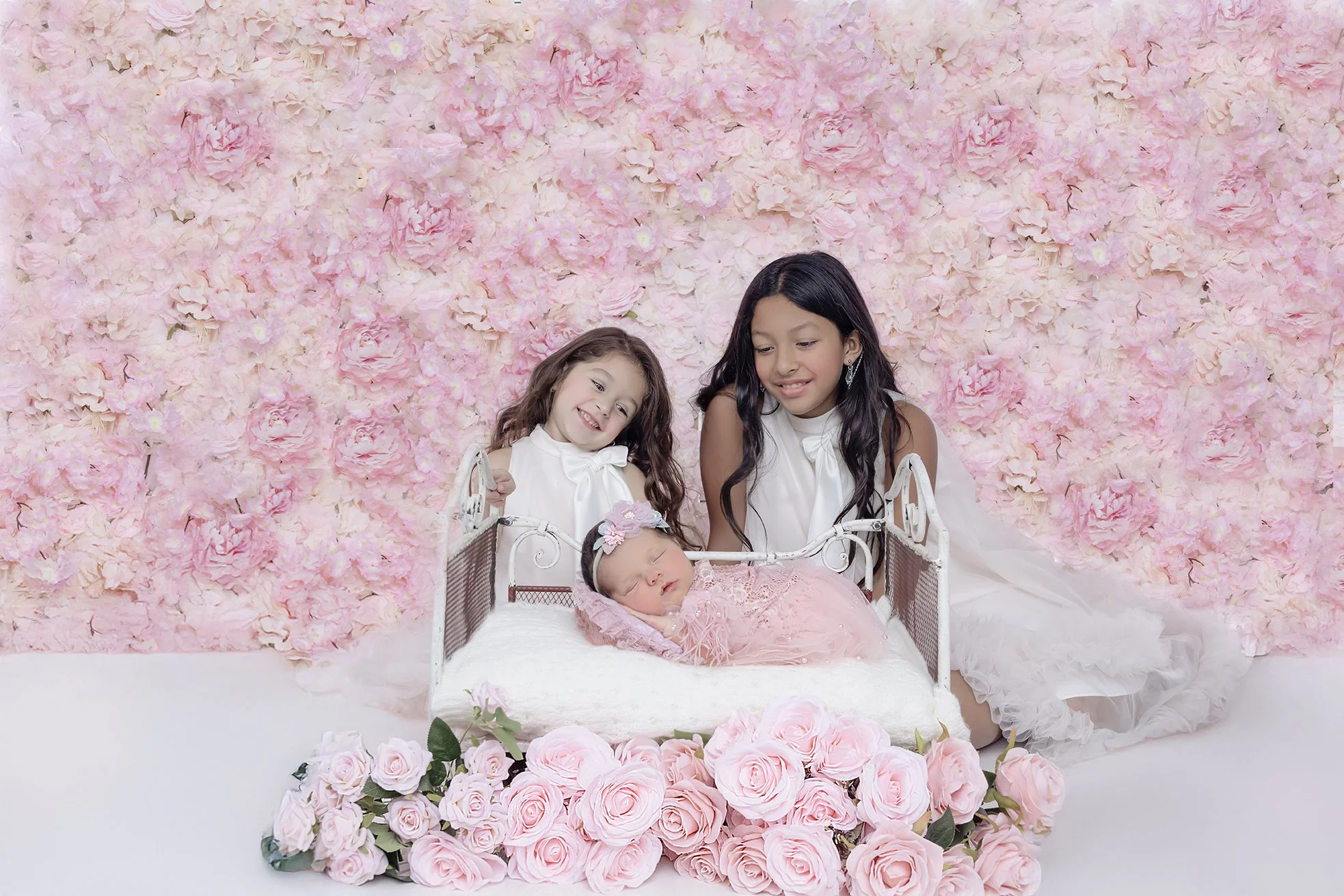 Two young girls and a newborn baby girl in a pink dress, with floral headband and earrings, on a small white bed amidst pink roses, with a pink flower wall background.