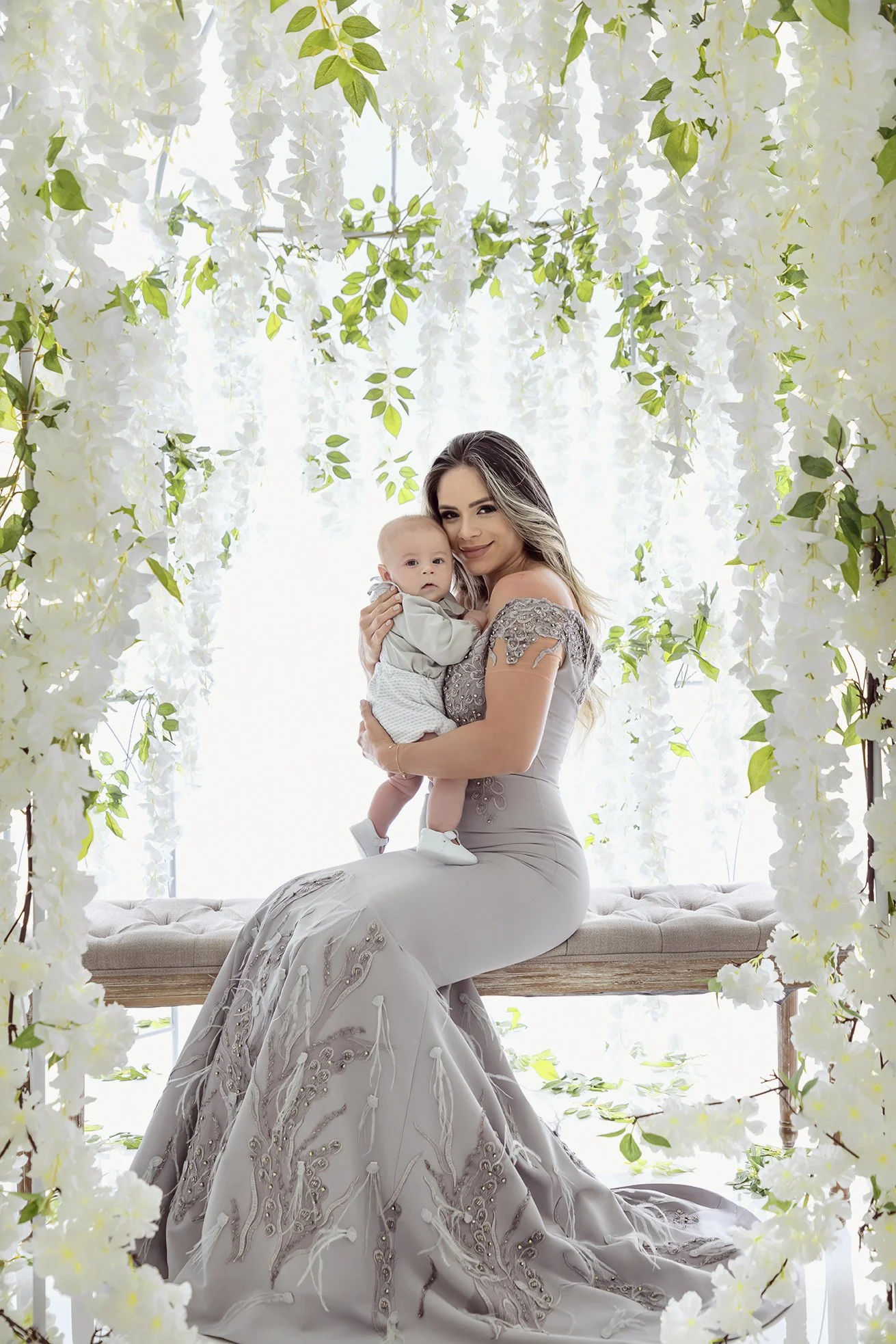 A woman dressed in a silver gown sitting on a wooden bench holding a young child surrounded by white floral decorations.