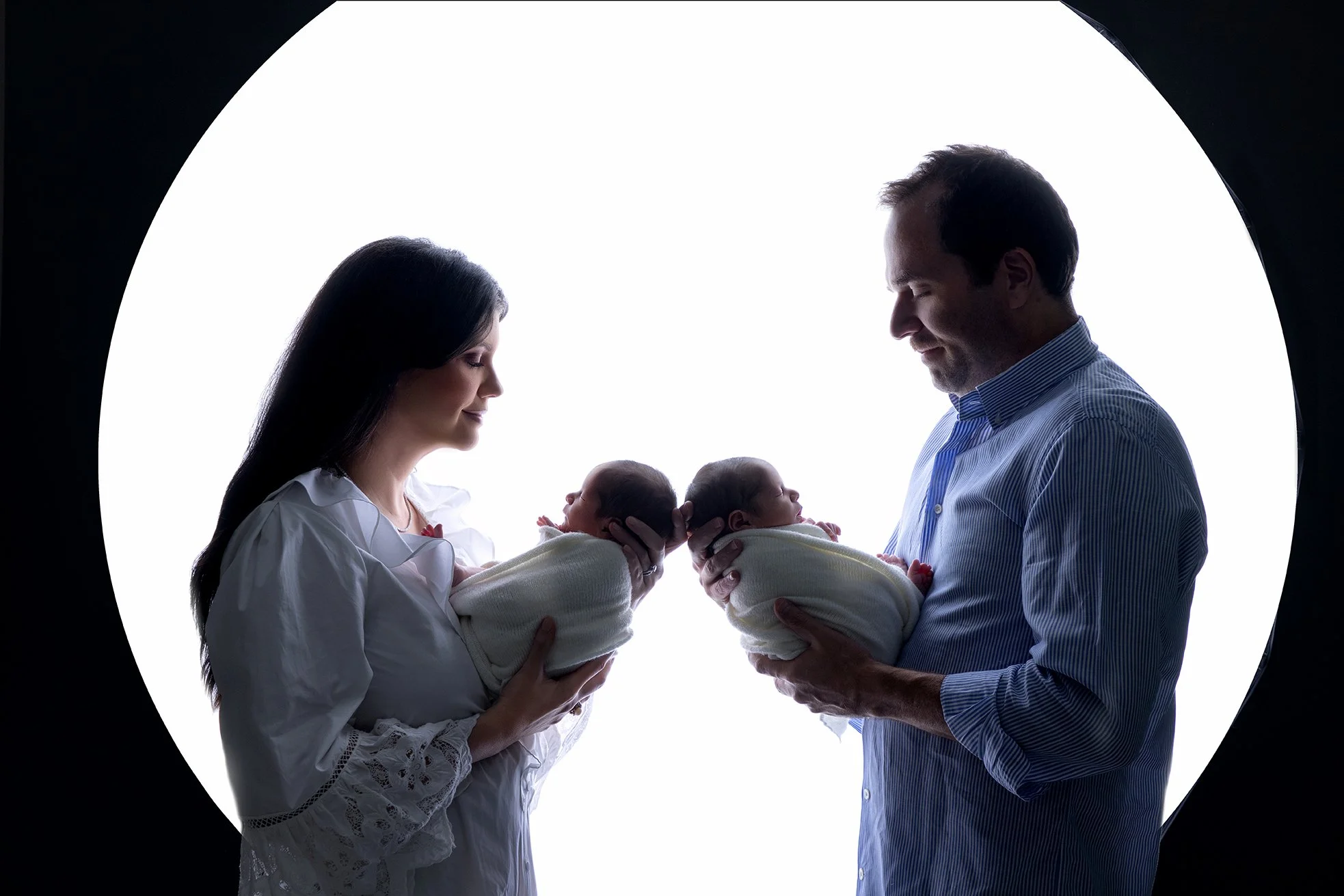 A man and woman holding newborn babies closely in silhouette against a bright, circular background.