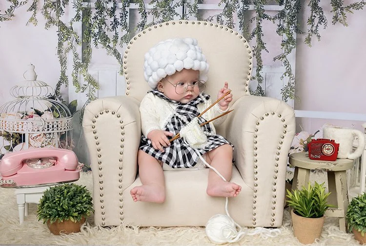 Young child dressed in a knitted hat, glasses, and a black-and-white checkered dress knitting with yarn and knitting needles while sitting on a beige armchair. The scene includes decorative greenery, a pink vintage phone, a small red radio, and potte
