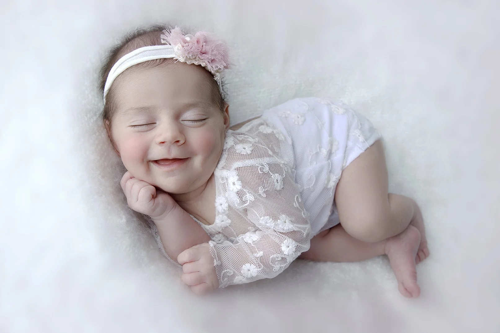 Smiling baby girl lying on her side on a soft, white surface with her eyes closed, wearing a pink headband with a flower, lace dress, and smiling with her hand under her chin.