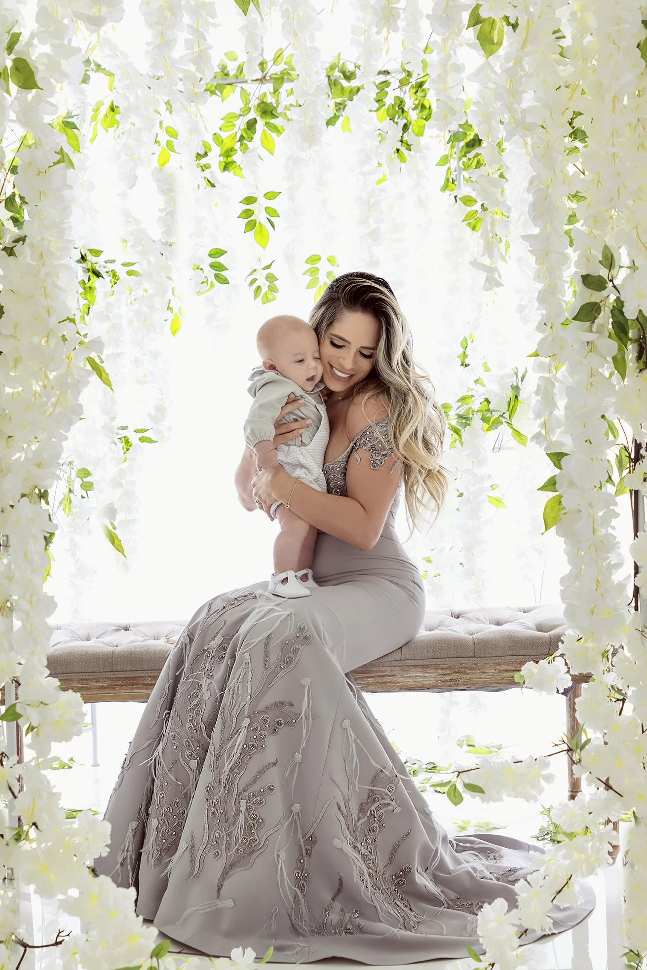 A woman in an elegant gown holding a baby surrounded by white flowers and green foliage.