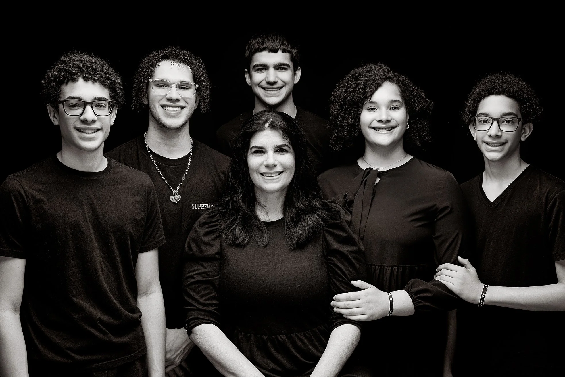 A woman and five teenagers posing together, all wearing black tops, against a dark background.
