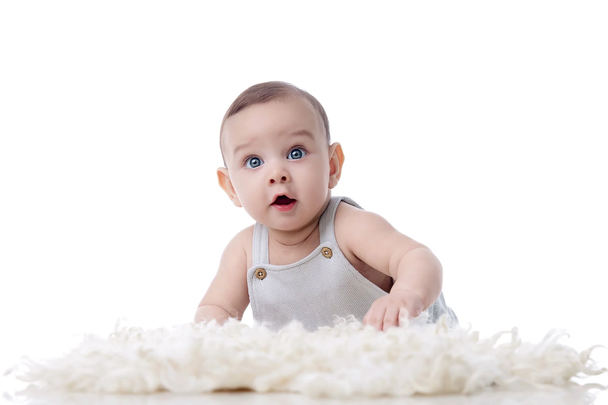 Baby with blue eyes in a sleeveless gray shirt, lying on a cream-colored fuzzy rug, looking at the camera with an expression of surprise.