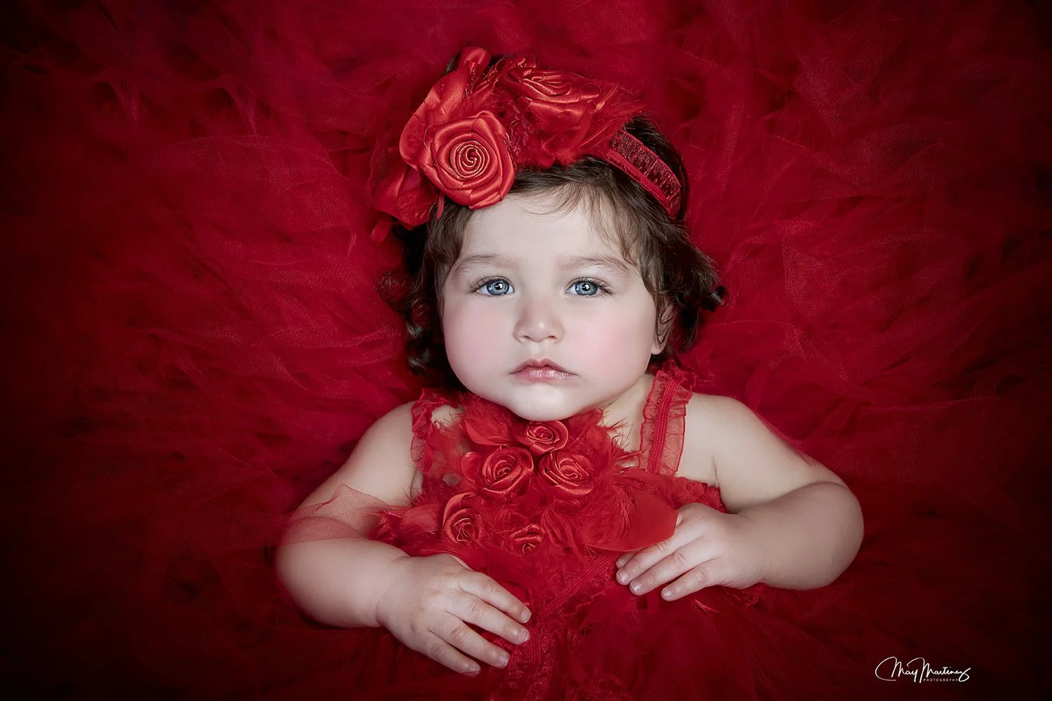 A young girl with blue eyes and brown curly hair lies on a red tulle background. She wears a red dress with rose and feather details, and a headband adorned with red roses.
