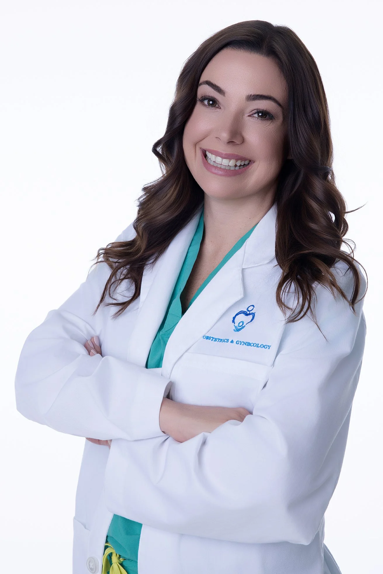 A woman with long wavy brown hair smiling, wearing a white medical coat with green scrubs underneath, arms crossed, against a plain white background.