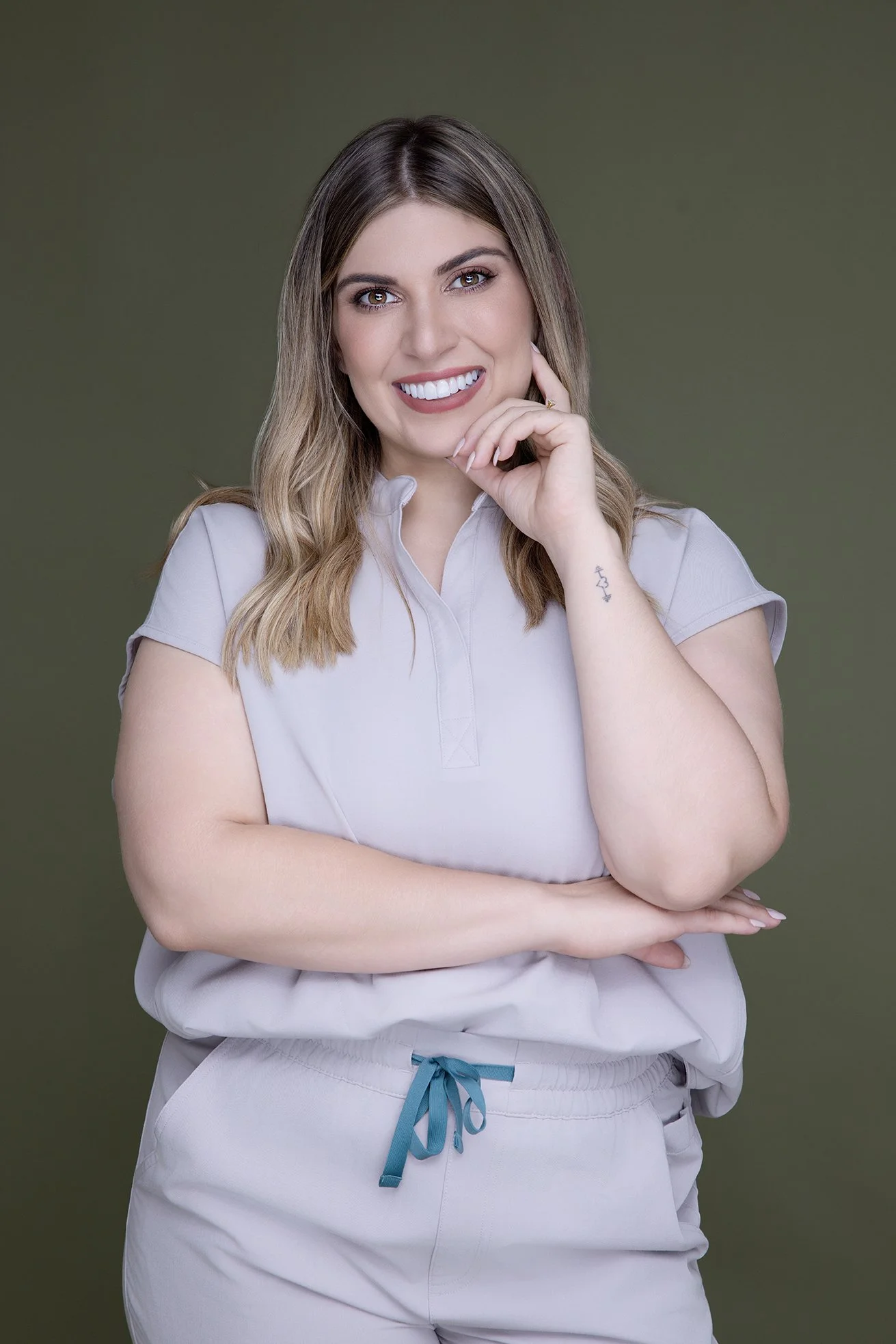 A woman with shoulder-length wavy hair, smiling, wearing a light gray short-sleeve top and matching shorts with a blue drawstring, posed with her left hand touching her chin and her right arm crossed over her waist, against a plain olive green backgr