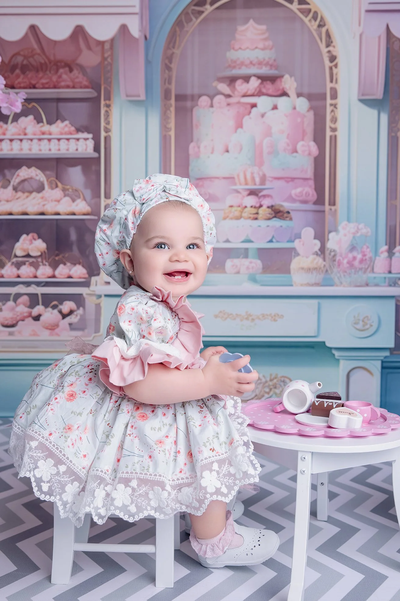 A baby girl sitting on a small white chair in front of a pink and blue dessert-themed backdrop, holding a toy teapot, with a pink table full of toy desserts.
