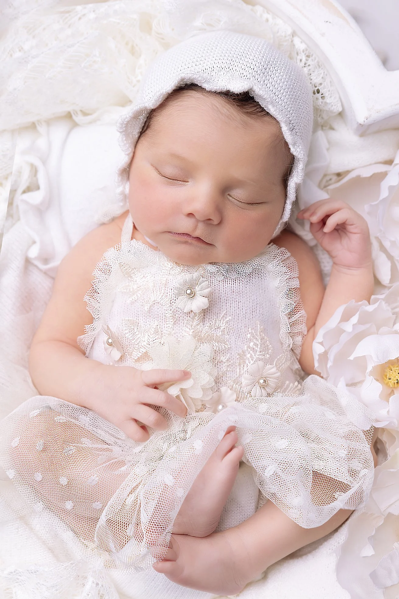 A sleeping baby girl with a headband, dressed in an off-white, lace outfit, surrounded by soft, white fabric and paper flowers