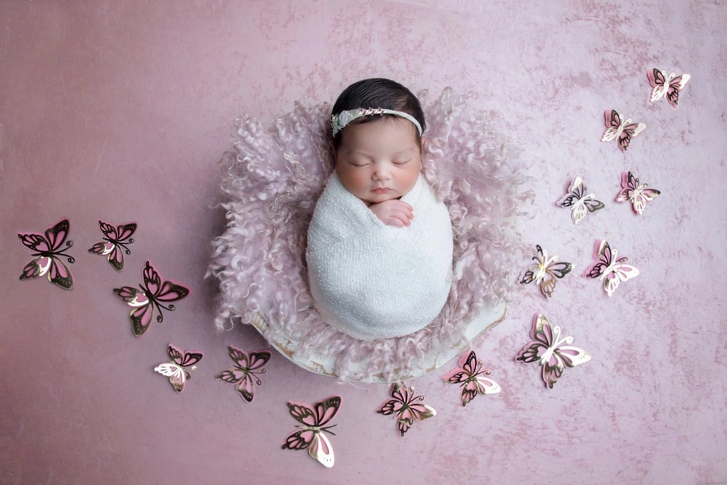 A newborn baby sleeping, wrapped in a white blanket, surrounded by pink curly fur, lying on a pink background with pink and white butterfly cutouts around.