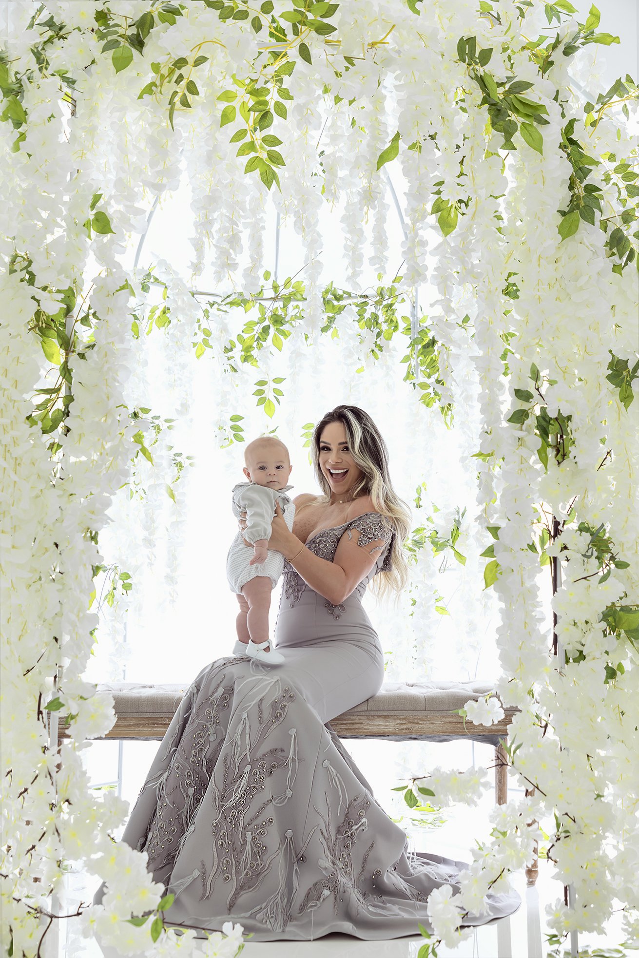 A woman in a silver gown sitting on a wooden bench, holding a baby dressed in light-colored clothes, under an arch of white flowers and green leaves.