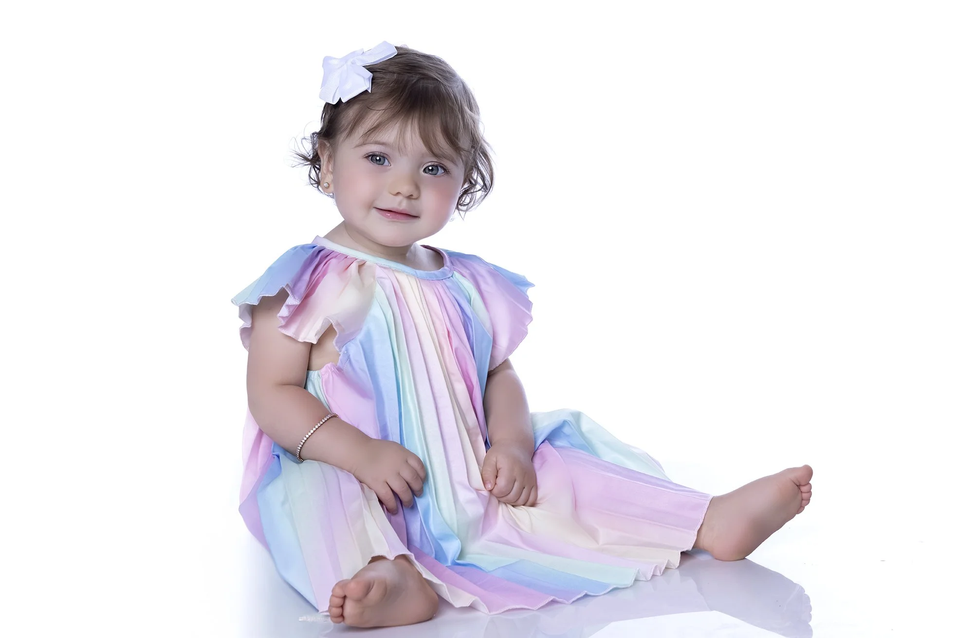 A young girl sitting on the floor, wearing a pastel rainbow-colored dress with ruffled sleeves and a white bow in her hair.
