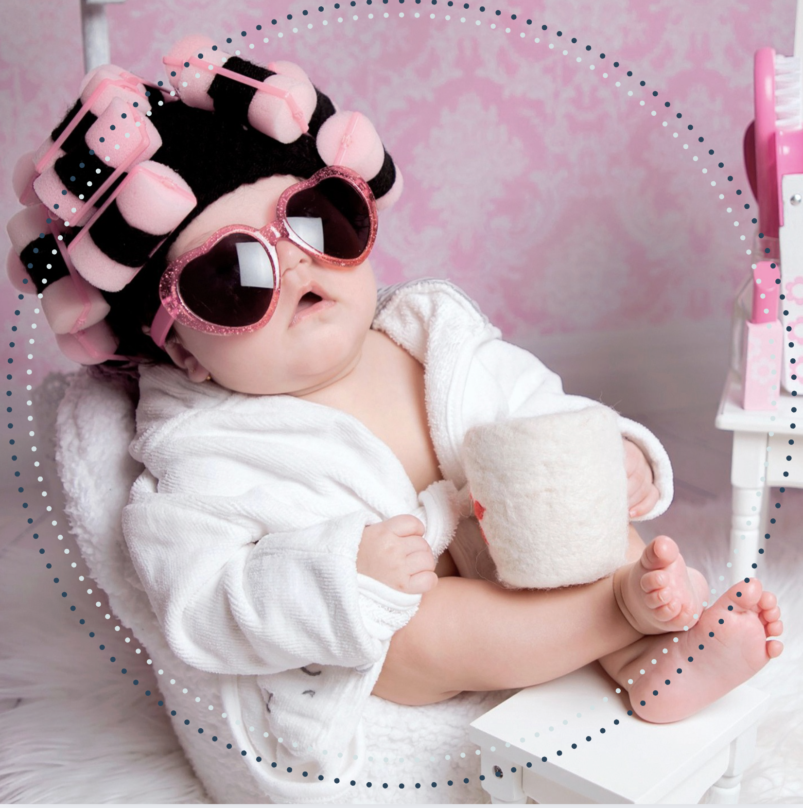 A baby wearing oversized pink heart-shaped sunglasses and pink hair rollers, sitting in a cozy white chair, holding a plush cup, against a pink background.