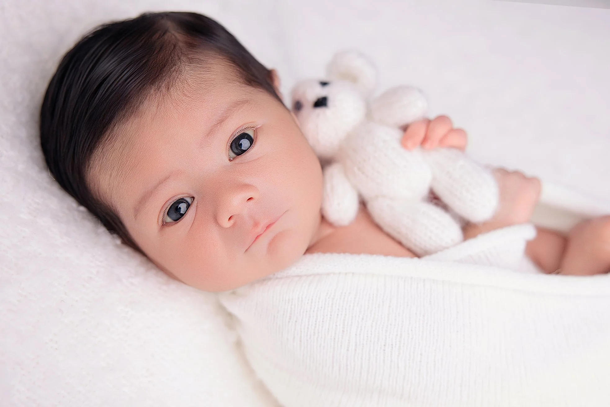 Close-up of a baby with dark hair lying on a soft white surface, holding a knitted white teddy bear.