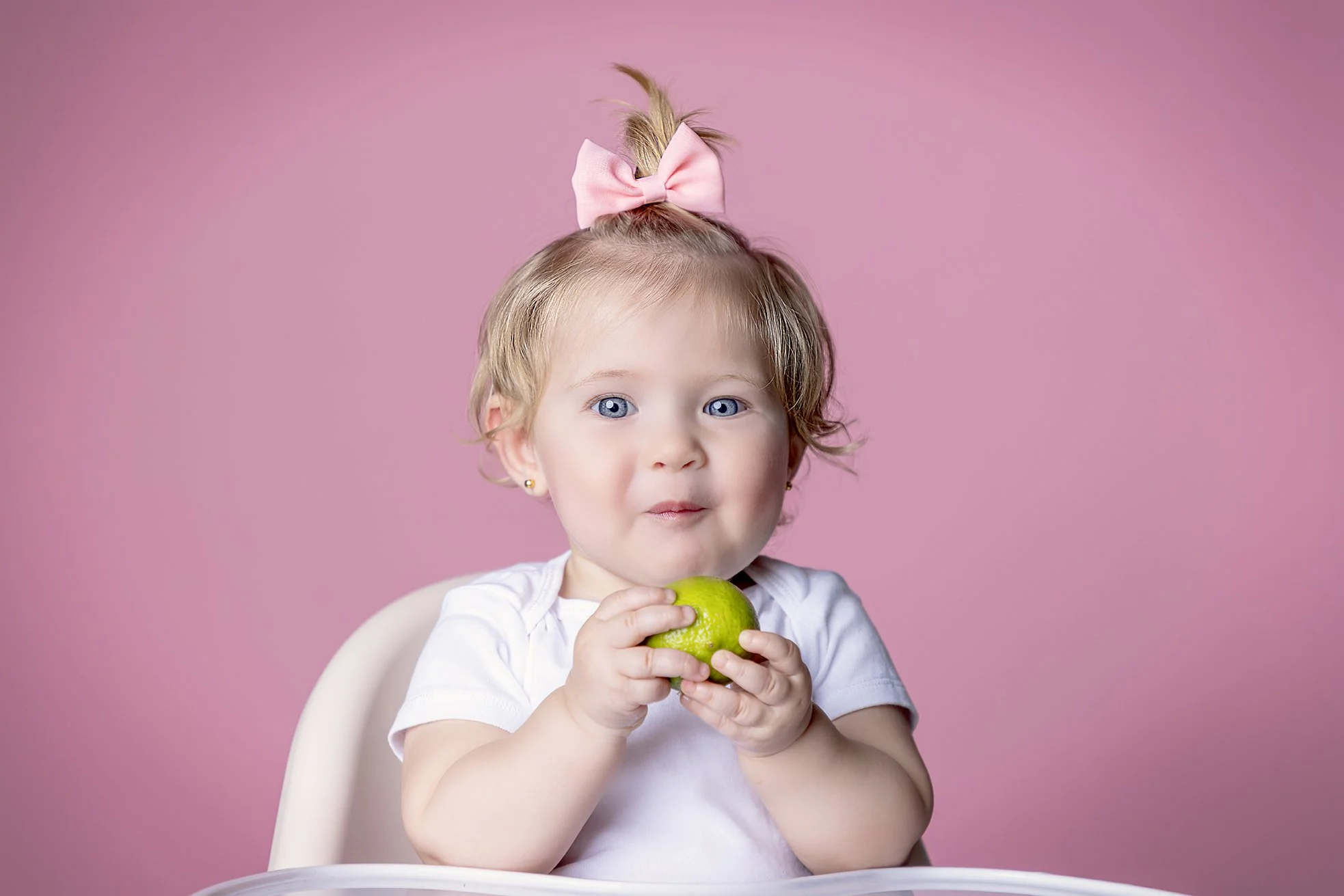 Young girl with blue eyes, blonde hair, pink bow, holding a green apple, sitting in a high chair against a pink background.