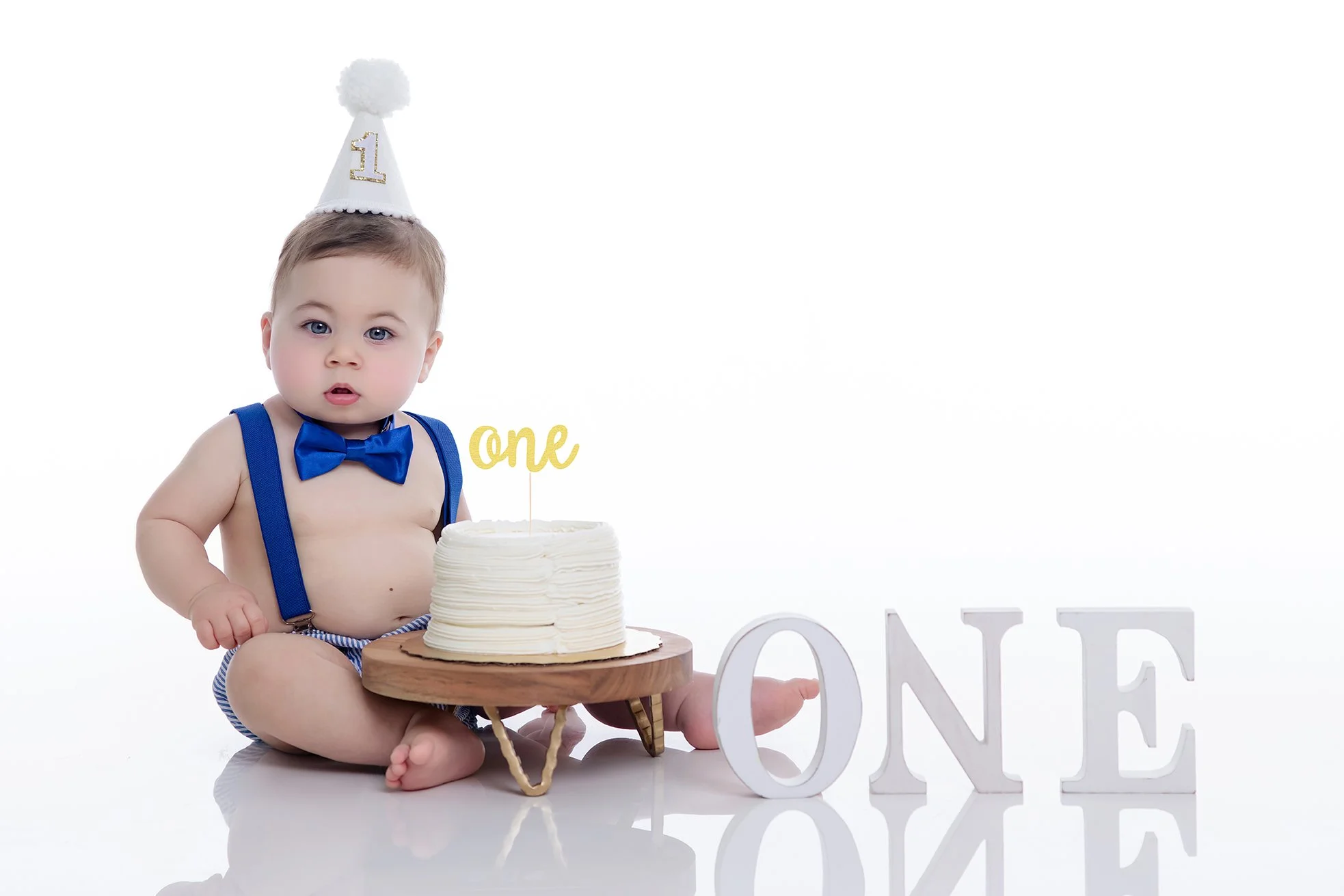 Baby boy celebrating a first birthday, wearing a birthday hat and a blue bow tie, sitting with a cake on a wooden stand, with 'one' written on a cake topper and large white letters spelling 'ONE' beside him.