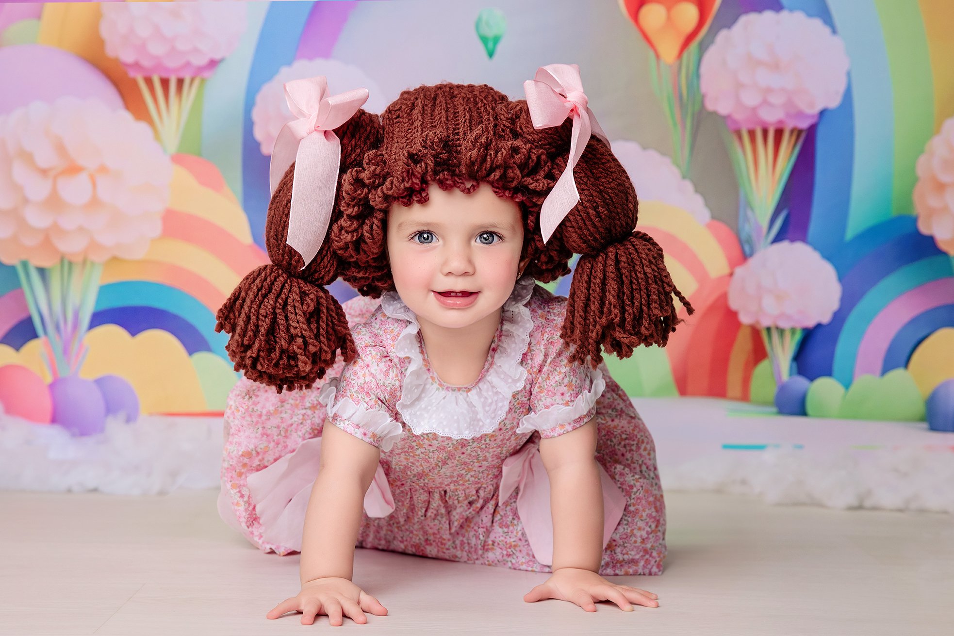 Young girl crawling on the floor in front of a colorful rainbow and cloud backdrop, wearing a pink floral dress and large brown yarn pigtails with pink bows.