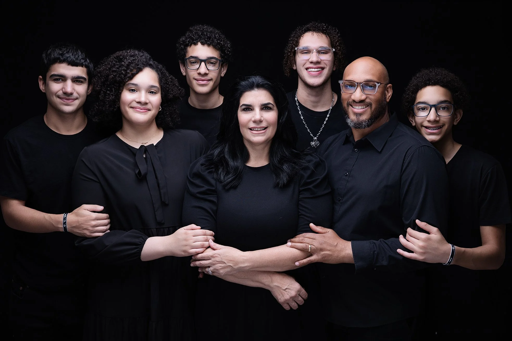 A diverse group of eight people, including adult man and woman and six young adults, standing together against a black background, all dressed in black and smiling.