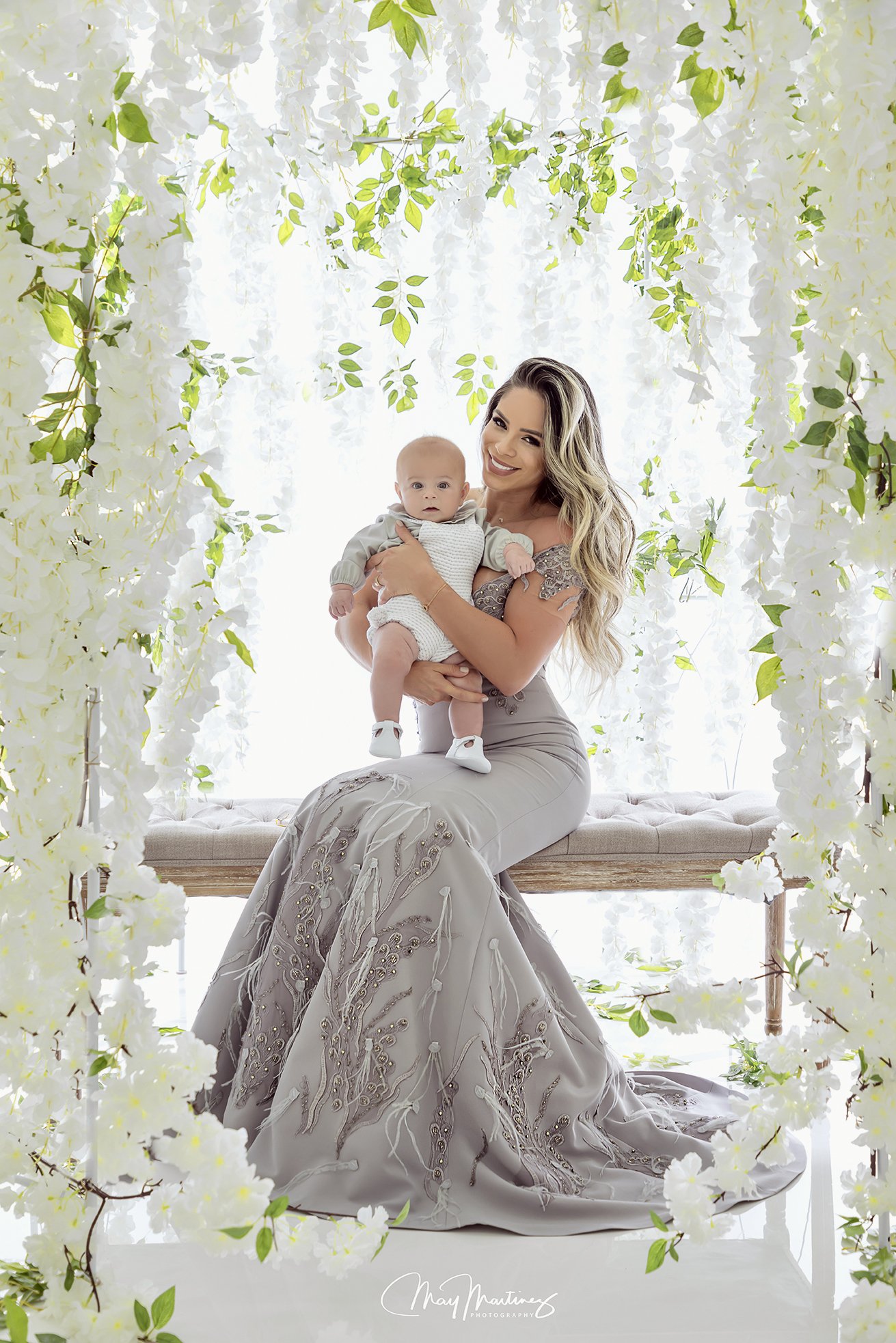 A woman in a gray, embroidered gown sitting on a bench holding a baby dressed in a light-colored outfit, surrounded by white flowers and green foliage in a bright setting.